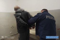 Na zdjęciu Policjant prowadzi zatrzymanego mężczyznę, którym ma skute ręce w kajdanki.