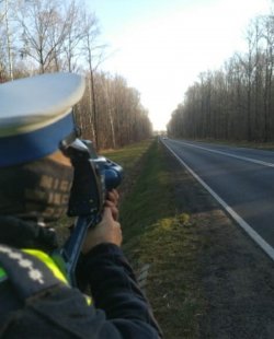 Na zdjęciu droga i policjant trzymający w ręku urządzenie mierzące prędkość nadjeżdżającego pojazdu.