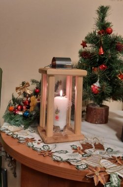Zdjęcie przedstawia Betlejemskie Światełko Pokoju, które stoi na pokrytym obrusem stole. W tle znajdują się ozdoby świąteczne.