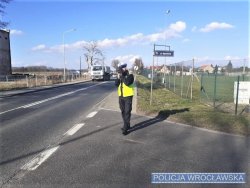 Zdjęcie przedstawia stojącego na na poboczu policjanta trzymającego w ręce przyrząd do pomiaru prędkości pojazdów. W tle widać jadące samochody.