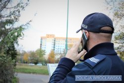 Na zdjęciu policjant stoi przed blokiem mieszkalnym i rozmawia przez telefon komórkowy z mieszkańcami na kwarantannie.