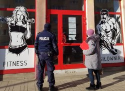 Na zdjęciu policjant z pracownicą sanepidu stoją przed budynkiem fitness i siłowni.