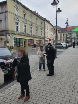 Na zdjęciu policjant i strażnik miejski rozmawiają na chodniku z seniorkami i przekazują im ulotki.