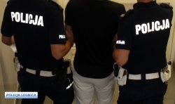 Na zdjęciu dwóch policjantów ruchu drogowego prowadzi zatrzymanego w czarnej koszulce i białych spodniach.