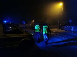 Umundurowani policjanci w czasie patrolu, w pobliżu przejścia dla pieszych. Zdjęcie zrobione nocą.