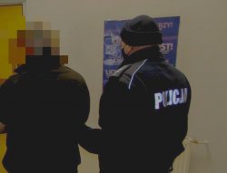 Na zdjęciu policjant w mundurze w maseczce ochronnej na twarzy prowadzi korytarzem zatrzymanego.