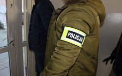 Na zdjęciu policjant w cywilnym ubraniu, z opaską na ręce z napisem policja prowadzi zatrzymanego mężczyznę korytarzem do aresztu.
