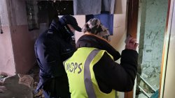 Umundurowany policjant wraz z przedstawicielką MOPS-u wychodzą  sprawdzają opuszczone budynki.