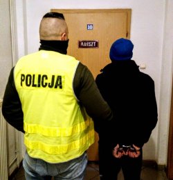 Na zdjęciu policjant w kamizelce odblaskowej z zatrzymanym mężczyzną., który na rękach ma założone kajdanki.