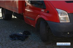 Zdjęcie lewego boku samochodu dostawczego koloru czerwonego patrząc z przodu pojazdu, stojącego na kostce brukowej przy drzwiach pasażera leży rozbite szkło i czarny foliowy worek.