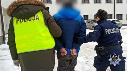 Na zdjęciu policjant i policjantka prowadzą zatrzymanego mężczyznę przez ośnieżony policyjny parking.