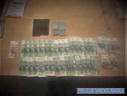 Na zdjęciu banknoty rozłożone na stole, kilkanaście sztuk o nominale po 100 złotych, 2 banknoty po 10 złotych, 2 banknoty po złotych, 1 banknoty 50 złotych i 1 banknot  200 złotych. Ponadto na stole znajdują się dwie wagi elektroniczne, oraz dwa telefony komórkowe.