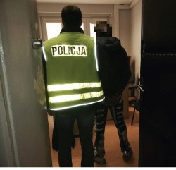 Na zdjęciu policjant wprowadza do pomieszczenia zatrzymanego mężczyznę z kajdankami na rękach.