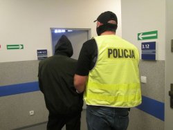 Na zdjęciu policjant prowadzi zatrzymanego mężczyznę przez korytarz komendy policji.
