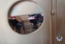 Na zdjęciu widok do pokoju przesłuchań przez wizjer do drzwi w którym policjant przesłuchuje zatrzymanego mężczyznę.