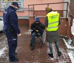 Przed budynkiem na chodniku stoi umundurowany policjant oraz pracownik ośrodka pomocy społecznej a na wózku siedzi osoba niepełnosprawna.