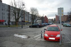 Na zdjęciu widać czerwony samochód zaparkowany na ulicy. W oddali inne pojazdy i okoliczne zabudowania.