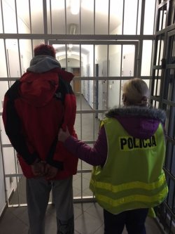 Policjantka w odblaskowej żółtej kamizelce z napisem POLICJA, trzyma za rękę podejrzanego mężczyznę. Oboje stoją tyłem przed pomieszczeniem z metalowymi kratami.
