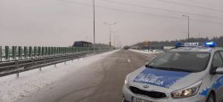 Na drodze zaparkowany jest policyjny radiowóz oznakowany. W tle widać samochody oraz śnieg.