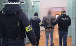 nie umundurowani policjanci z opaskami z napisem policja prowadzą mężczyznę korytarzem w budynku komendy