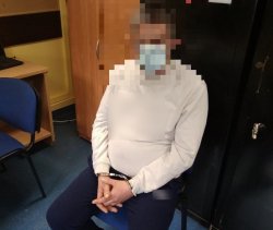 siedzący na krześle zatrzymany mężczyzna z kajdankami na rękach, w białej bluzie i maseczce na ustach
