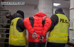 policjanci w kamizelkach prowadzą mężczyznę w czerwonej kurtce i kajdankach