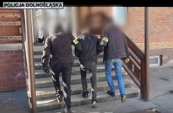nieumundurowani policjanci z opaskami na rękach prowadzą zatrzymanego mężczyznę