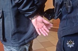 Na zdjęciu widać osobę w niebieskiej kurtce, która trzyma ręce z tyłu, widać również fragment sylwetki policjanta, który zakłada na ręce tej osoby kajdanki