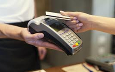 Na zdjęciu widać trzymany w rękach terminal płatniczy oraz rękę przykładającą do niego kartę bankomatową.