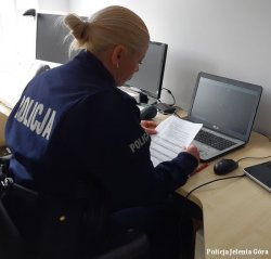na zdjęciu widać policjantkę siedzącą przy biurku przy komputerze i prowadzącą zajęcia zdalne