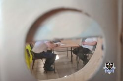 na zdjęciu widać przez wizjer siedzących przy biurku mężczyzn podczas przesłuchania, jeden to nieumundurowany policjant z kamizelką odblaskową powieszoną na oparciu krzesła