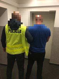 Na zdjęciu widać nieumundurowanego policjanta w kamizelce odblaskowej z napisem policja trzymającego pod rękę zatrzymanego, który ma założone kajdanki z przodu.