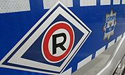 Na zdjęciu widać emblemat policyjny zamieszczony na drzwiach radiowozu. Emblemat to R w czerwonym okręgu wpisane w granatowy romb z białą obwolutą.