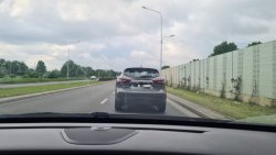 Na zdjęciu widać samochód osobowy jadący po jezdni. Auto widać z wnętrza innego pojazdu, przez jego przednią szybę.
