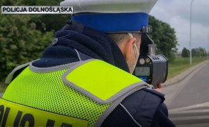na zdjęciu widać policjanta w kamizelce, stojącego na poboczu, patrzącego przez rejestrator prędkości
