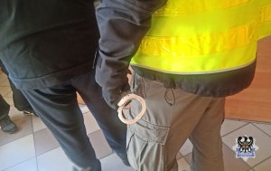 Funkcjonariusz policji w kamizelce odblaskowej stoi obok zatrzymanego mężczyzny z kajdankami w ręku.
