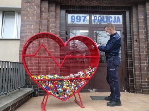 Umundurowany policjant stoi przed budynkiem jednostki policyjnej i wrzuca nakrętki do specjalnego, metalowego pojemnika w kształcie serca.