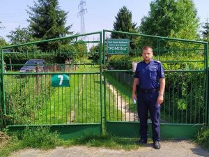 Zdjęcie przedstawia umundurowanego policjanta, który stoi przed bramą ogrodów działkowych