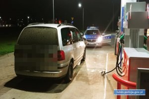 Zdjęcie przedstawia stacje benzynową, na której stoi srebrny samochód osobowy a przed nim radiowóz