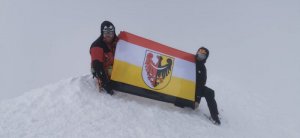 Na zdjęciu widać dwóch mężczyzn na śniegu, którzy trzymają flagę