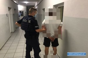 Zdjęcie przedstawia policjanta, który stoi na korytarzu z mężczyzną, który ma założone kajdanki na ręce trzymane z tyłu